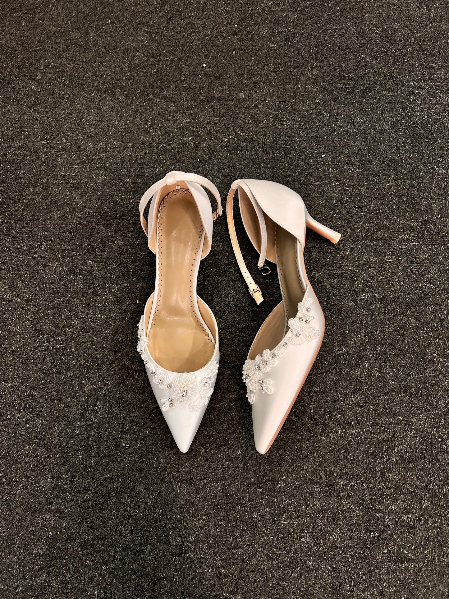 They are adorned with hand-placed lace floral appliqués that wrap elegantly around the outer edge of the pointed toe. Accented with micro-pearls and shimmering crystals, these heels offer a romantic, multidimensional look for the modern bride