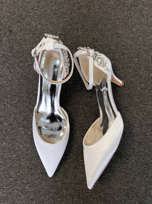 A pair of white pointed-toe high heels with silver metallic inner soles and crystal-embellished ankle straps, displayed on a dark gray textured surface.