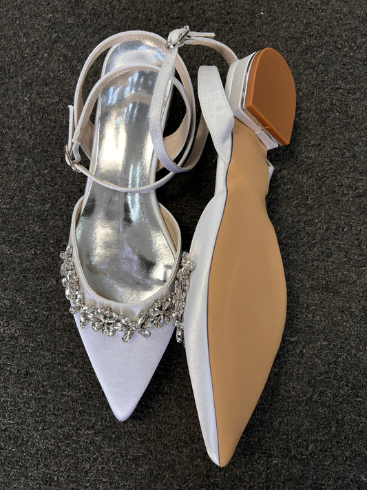 A pair of white satin pointed-toe flats with silver rhinestone embellishments on the toe, shown on a dark gray textured carpet. One shoe is oriented to show the white strap and silver insole, while the other shows the beige outsole and brown heel.