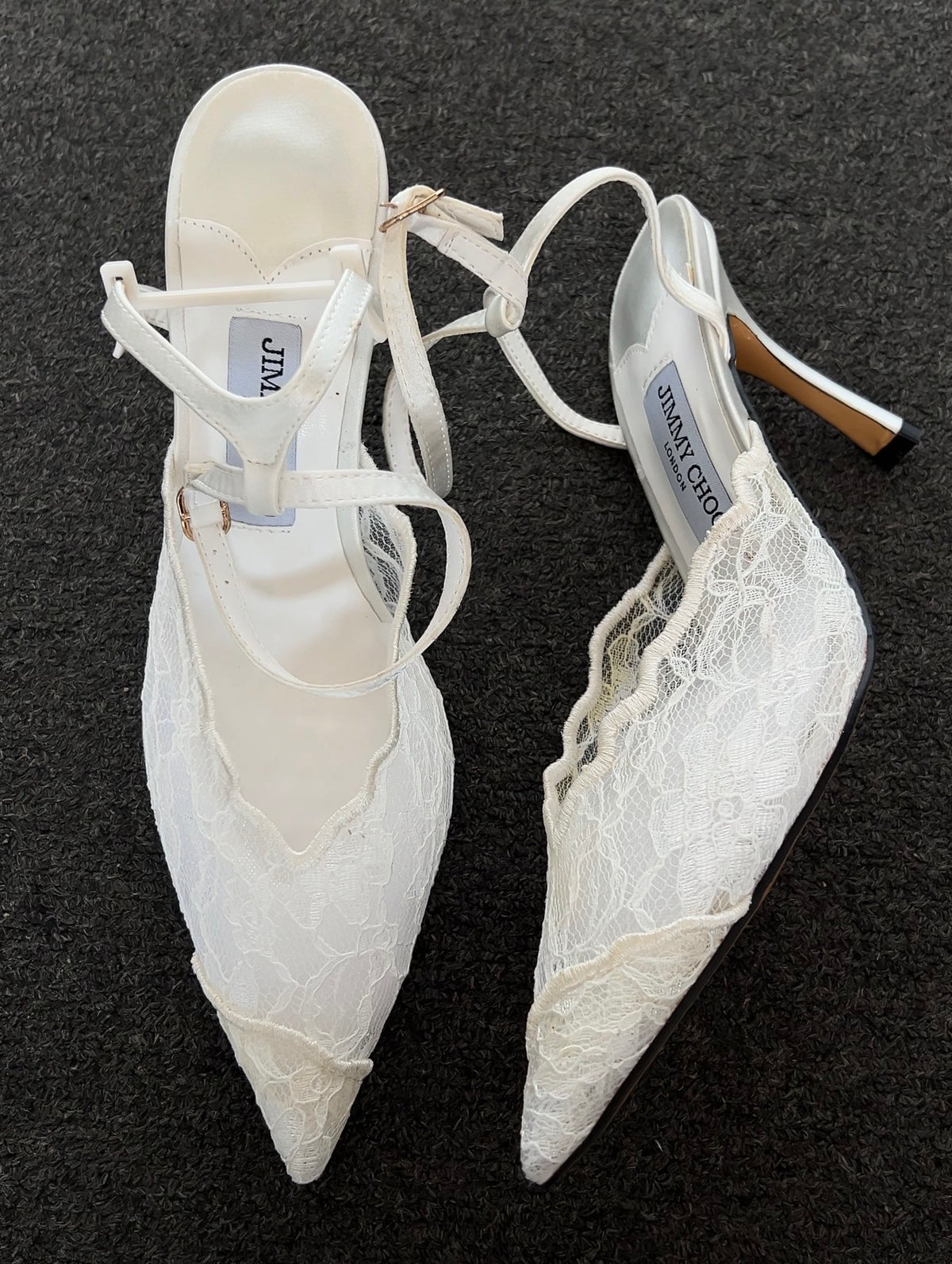 A pair of white pointed-toe slingback heels with lace overlays and thin wraparound straps, displayed on a dark textured background. The brand name 'JIMMY CHOO' is visible on the insole labels.