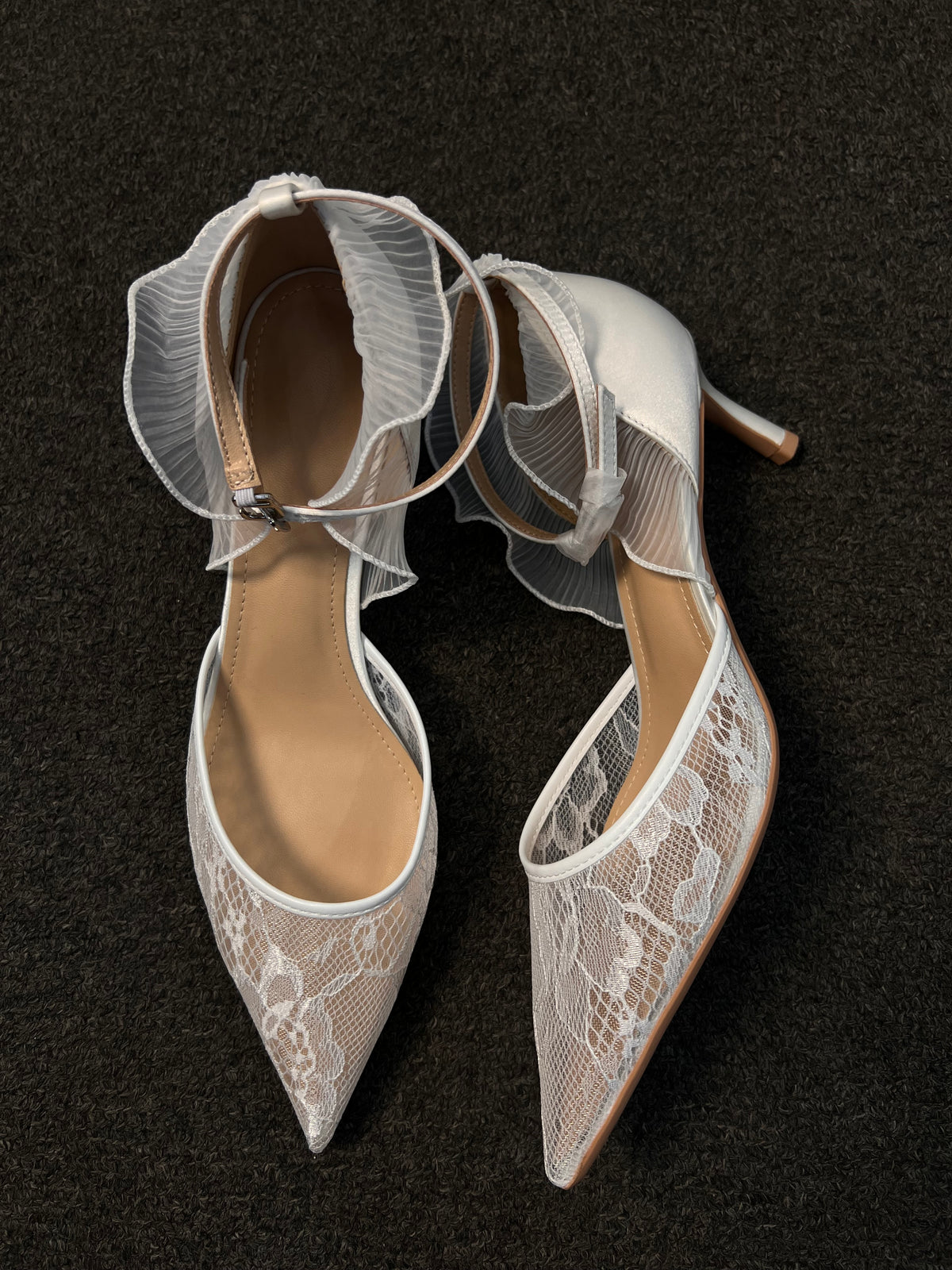 A pair of pointed-toe high heels in white lace and satin, featuring sheer ruffled ankle straps.