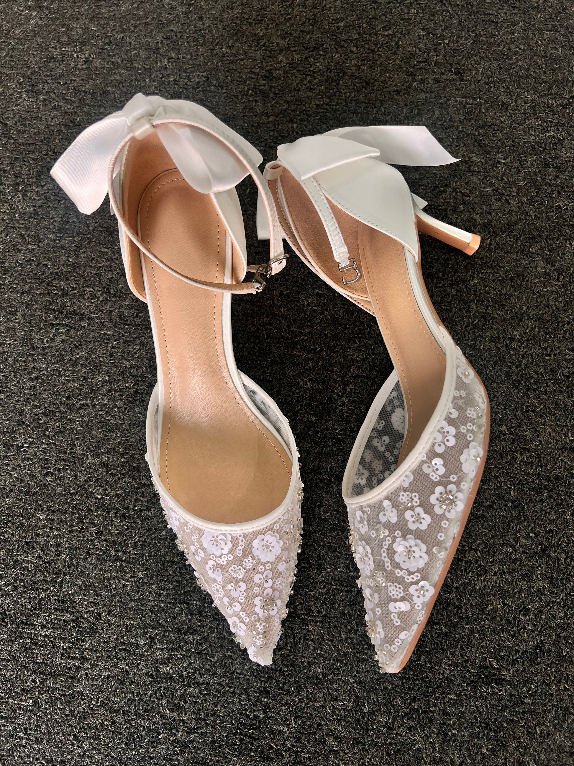Pair of white wedding shoes with lace detailing . The design is beautifully balanced by a smooth satin heel and an oversized satin bow at the back, adding a dramatic yet feminine finishing touch.